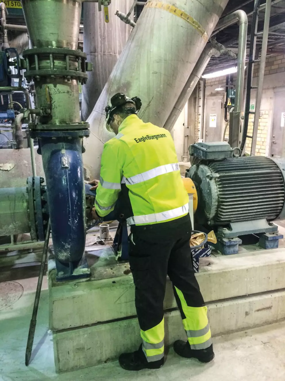 A technician wearing a high-visibility jacket with the EagleBurgmann logo is working on industrial machinery in a factory setting, surrounded by large metal pipes and equipment.