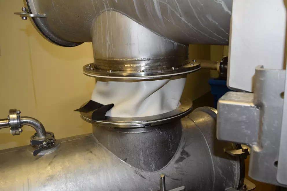 The image shows a metal piping system with a deformed white expansion joint at the junction, secured with black straps. The pipes are made of stainless steel.