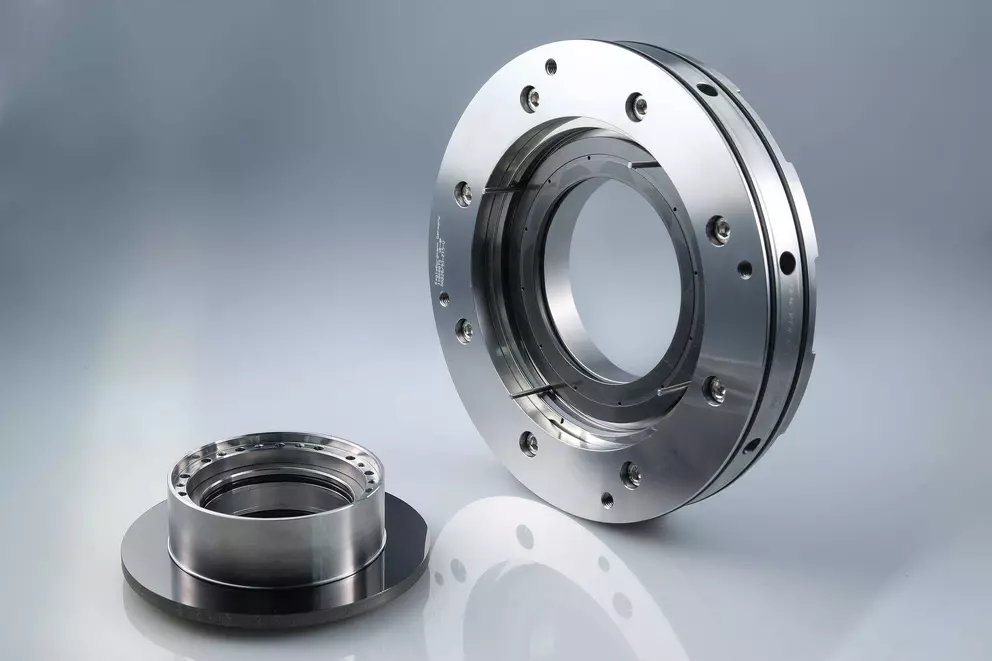 A studio photograph of two precision machined metal components, likely parts of a large mechanical seal or bearing: a large, bolted ring on the right, and a smaller flanged component resting on the left.
