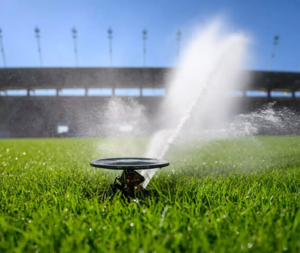 Automatisches Sprinklersystem, das bei hellem Sonnenlicht grünes Gras auf einem Sportplatz bewässert.