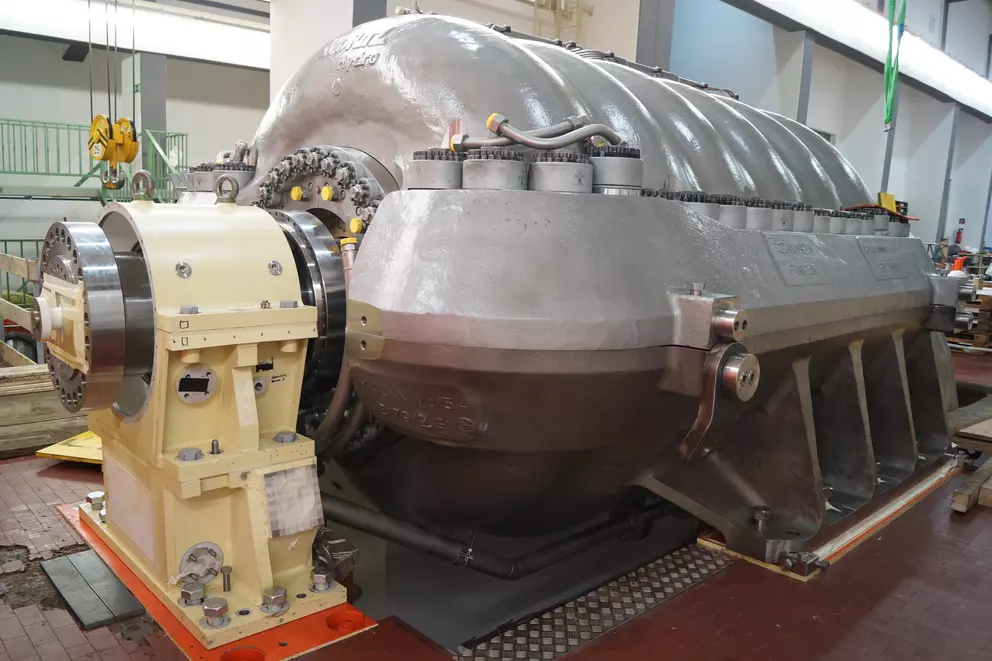 A close-up of a large industrial machine, likely a turbine or compressor, featuring a ribbed gray casing and a large tan-colored end section bolted to the left, on the floor of a factory hall.