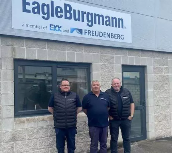 Three employees stand in front of the entrance to an EagleBurgmann building beneath a company sign featuring the EagleBurgmann, EKK, and Freudenberg logos.
