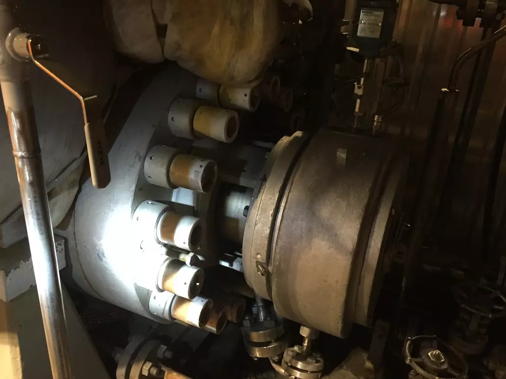 A close-up, dark photo of a heavily bolted industrial machine section, likely a turbine or large pump, showing a row of large bolts with ceramic or pale rings around them, partially illuminated by a bright spot of light.