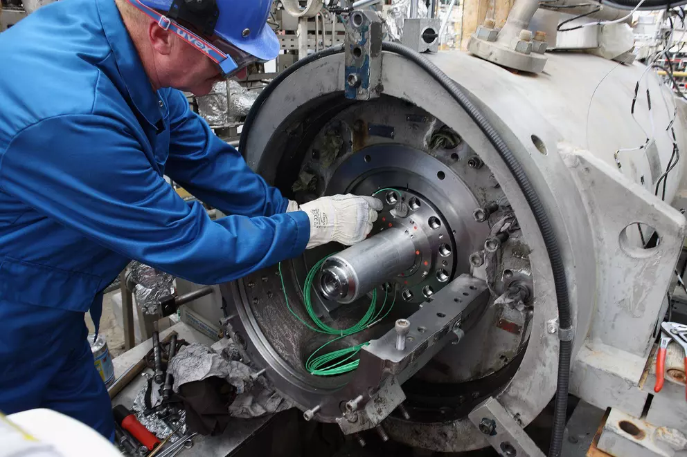 Ein Techniker in blauer Uniform, Schutzhelm und Schutzbrille installiert ein großes Bauteil oder eine Welle in einer komplexen Industriemaschine, wahrscheinlich einer Turbine oder einem Kompressor.