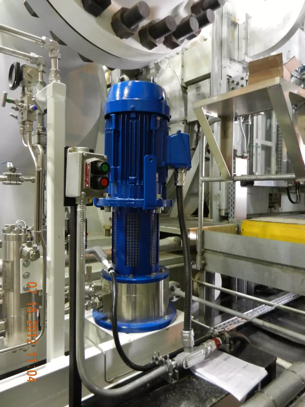A large blue industrial electric motor connected to a vertical silver pump assembly in a processing facility, with numerous pipes and control boxes visible. The time and date "07 18 2012 11:04" are stamped on the bottom left.