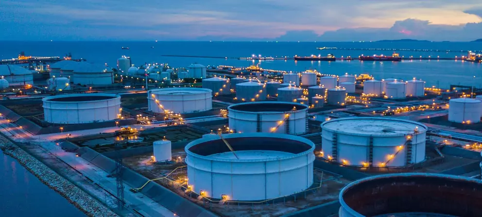 Aerial, twilight view of a large oil storage terminal on a coastline with numerous white cylindrical tanks and ships in the background.
