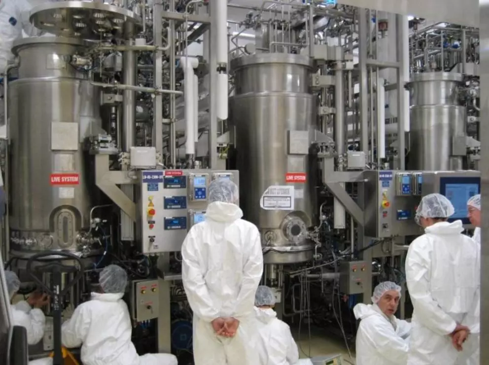 Workers in full-body cleanroom suits and hairnets operate and inspect large stainless steel bioreactors or fermenters inside a sterile pharmaceutical or biotechnology manufacturing facility.