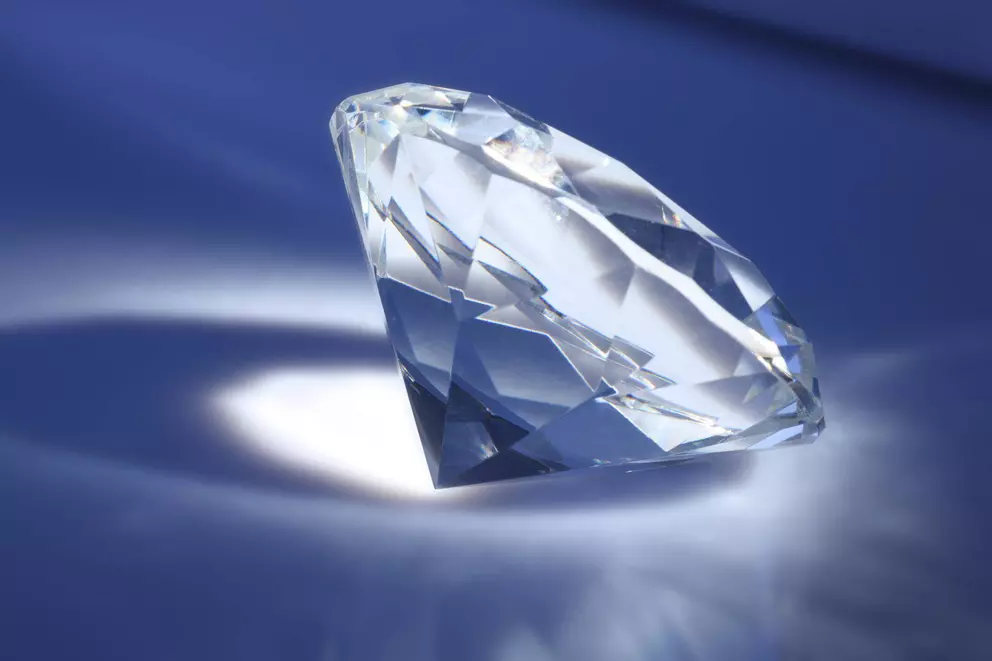 A single, brilliant-cut, faceted clear diamond or gem resting on a deep blue surface, illuminated by a spotlight that creates a bright reflection and sharp shadow.